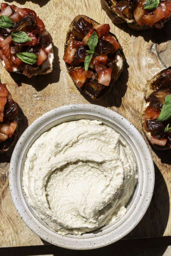 A bowl of vegan cashew cheese on a wooden cutting board with Date & Strawberry Crostini around the bowl.