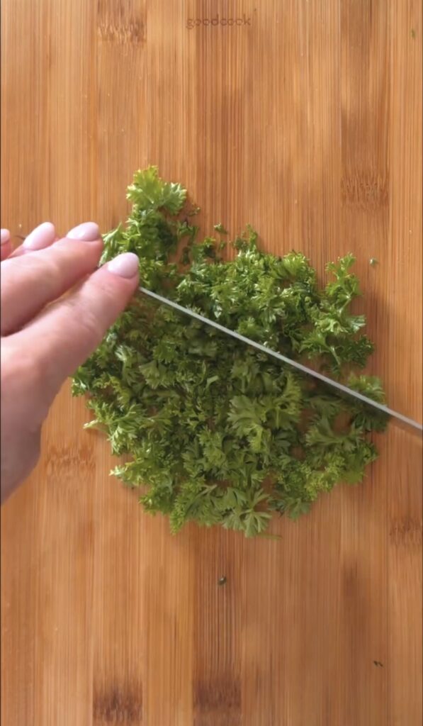 action shot mincing the fresh parsley with a knife on a wooden cutting board.