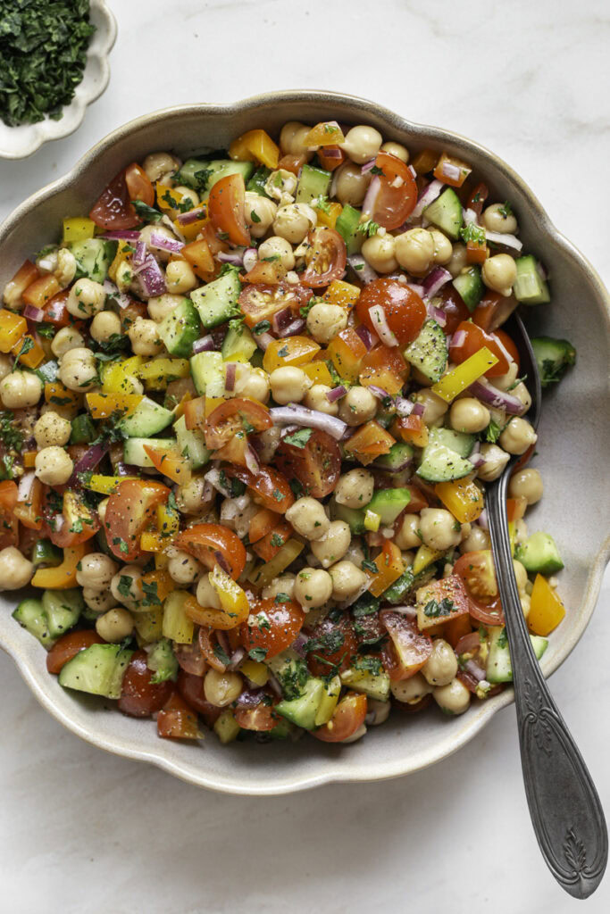 a bowl of chickpea salad with a serving spoon and a small bowl of chopped parsley above it.