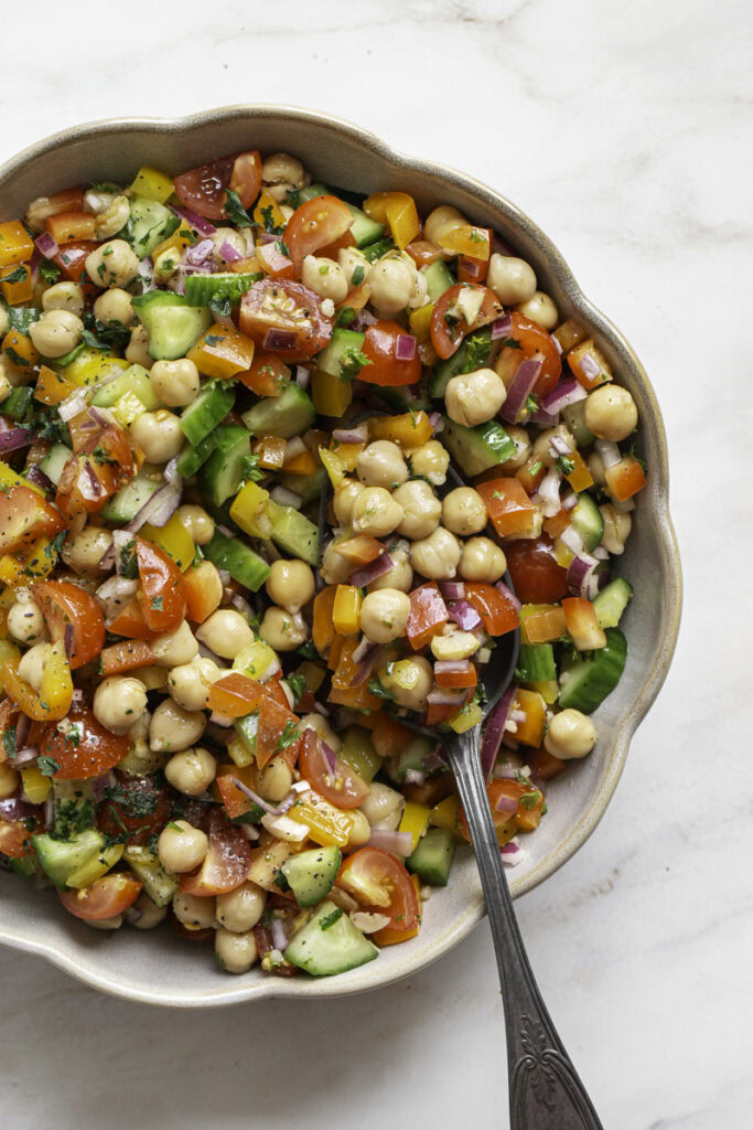 a bowl of chickpea salad with a serving spoon and a small bowl of chopped parsley above it.