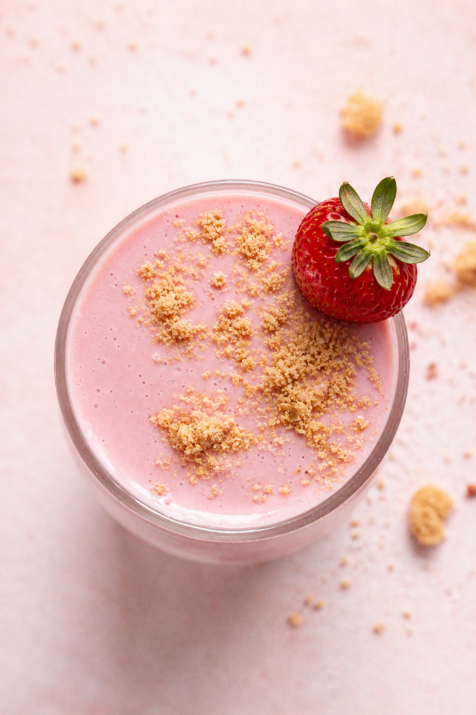 an overhead shot of a glass of strawberry cheesecake smoothie with graham cracker crumbs and a fresh strawberry on the rim of the glass.