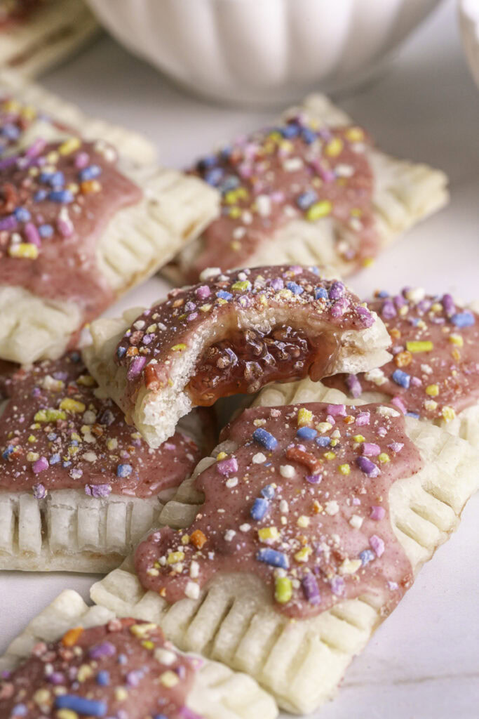 3/4 image of mini strawberry chia pop tarts piled on top of one another, the top one with a bite taken out of it to reveal the strawberry chia jam filling.
