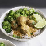 a bowl of rice topped with coconut curry meatballs, steamed broccoli, chopped cilantro served with lime wedges.