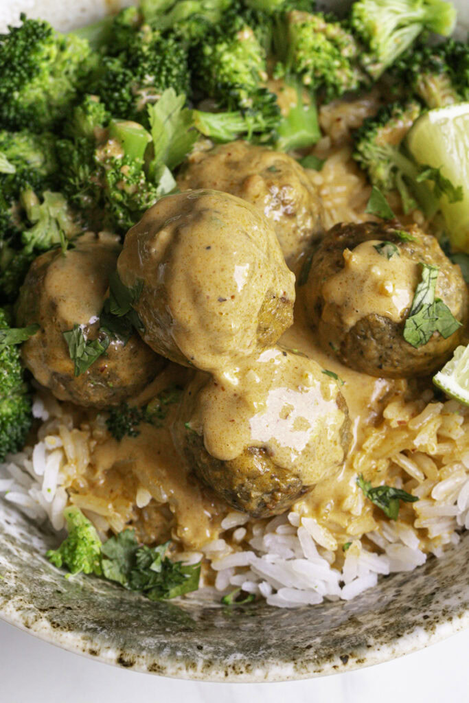 close up overhead shot of a bowl of rice topped with coconut curry meatballs, steamed broccoli, chopped cilantro served with lime wedges.