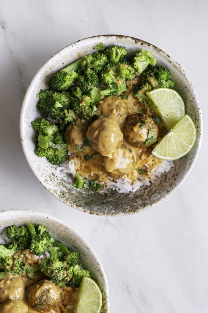 overhead shot of a bowl of rice topped with coconut curry meatballs, steamed broccoli, chopped cilantro served with lime wedges.