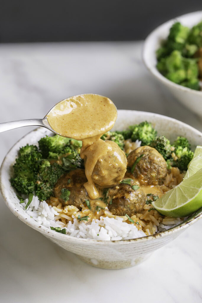 action shot drizzling coconut curry sauce over the meatballs and rice in a bowl.