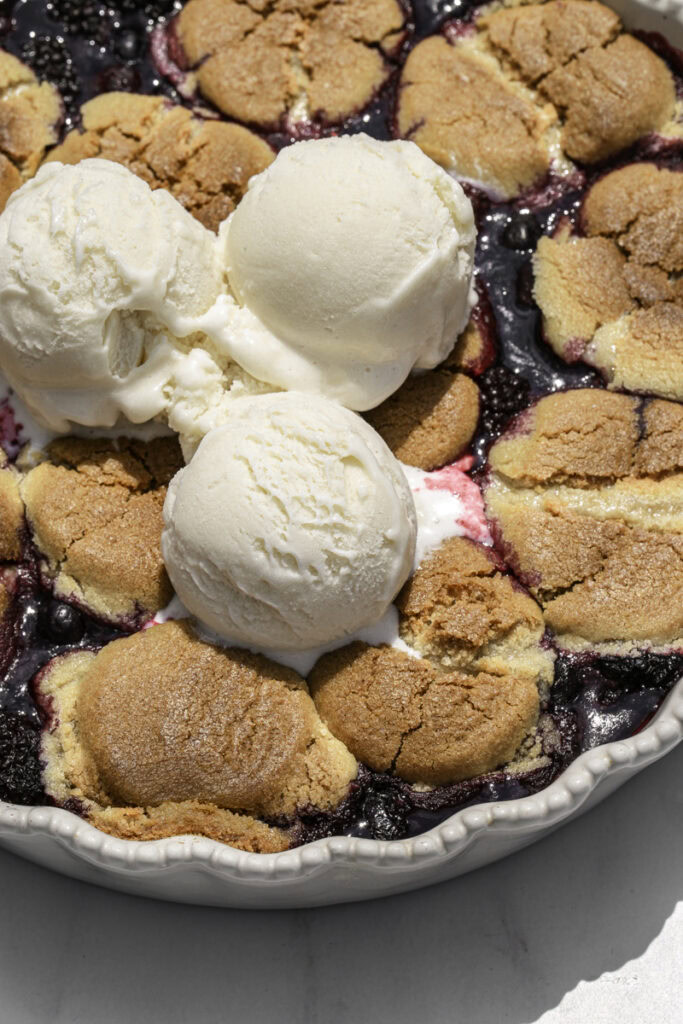 close up of baked cookie berry cobbler with 3 scoops of vanilla ice cream on top.