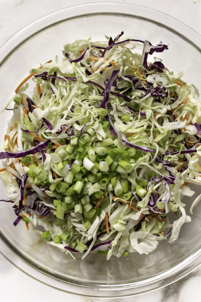 sliced green onions added to a large glass bowl with the shredded coleslaw mix.