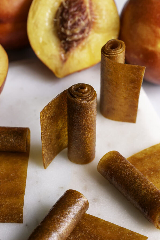rolled up nectarine fruit leather, some rolls standing vertically, some laying down. Fresh nectarines in the background.