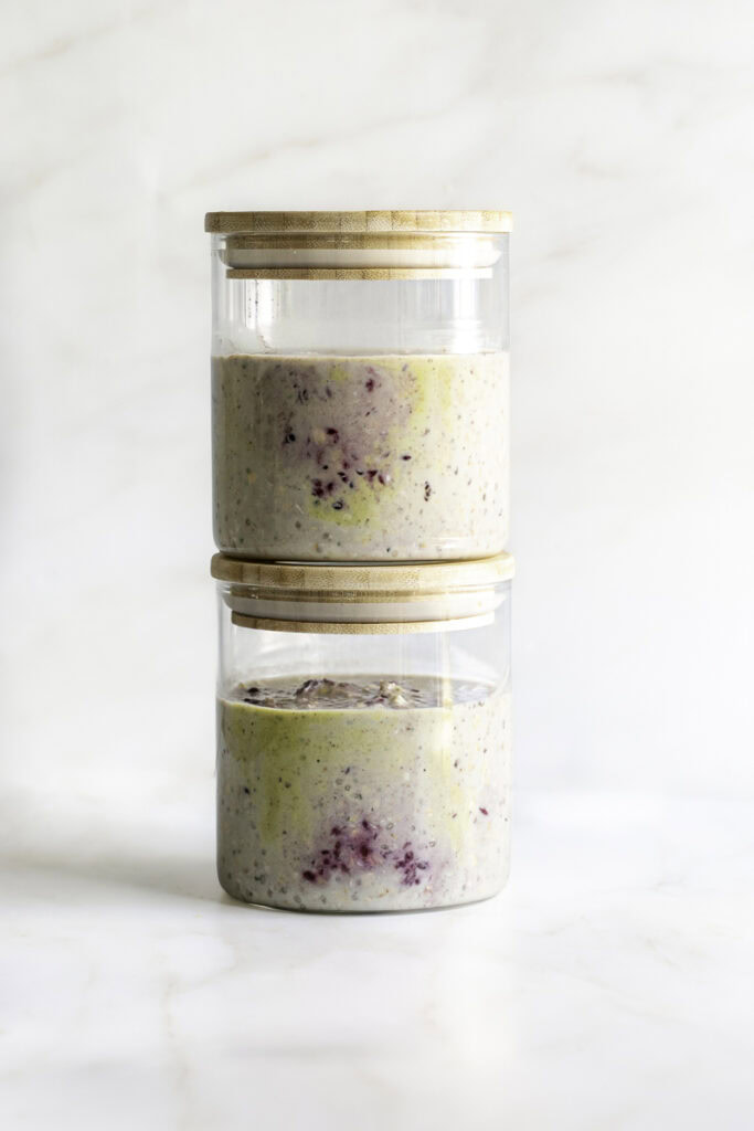 two jars of blackberry pistachio overnight oats with lids stacked on top of one another.