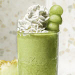 close up of a glass of honeydew green tea topped with whipped cream and a skewer of honeydew balls.