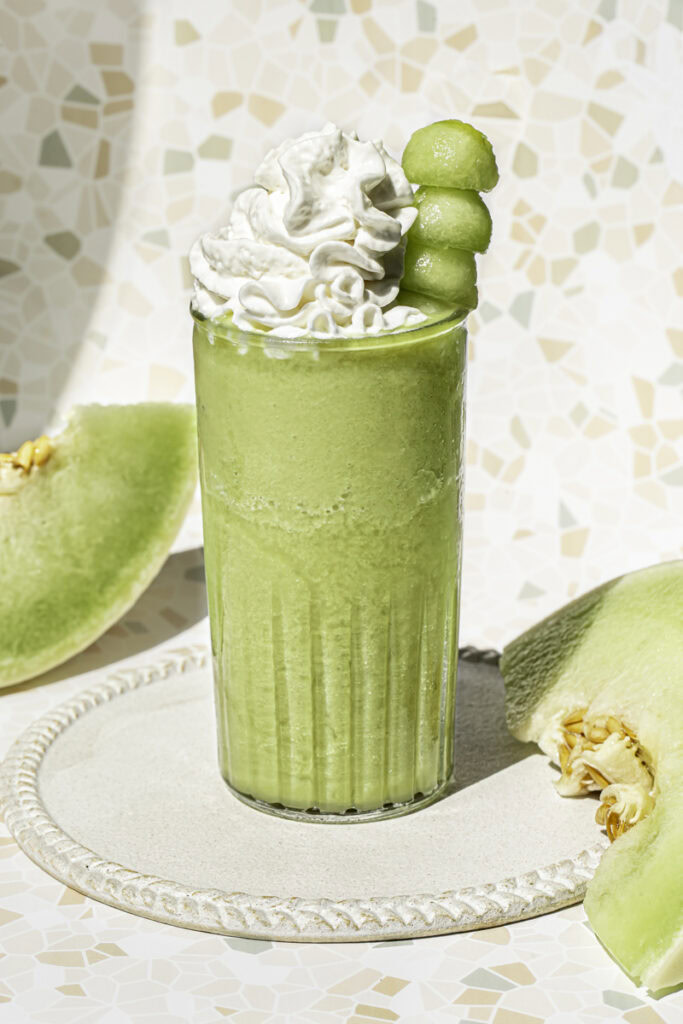 a glass of honeydew green tea topped with whipped cream and a skewer of honeydew balls and honeydew slices in the background.