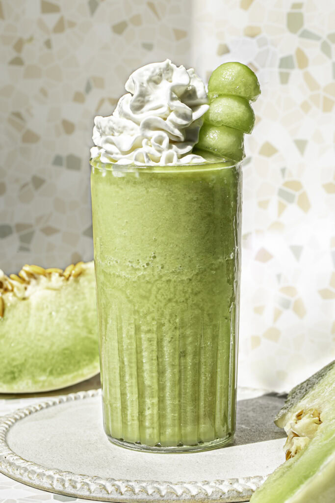 a glass of honeydew green tea topped with whipped cream and a skewer of honeydew balls.