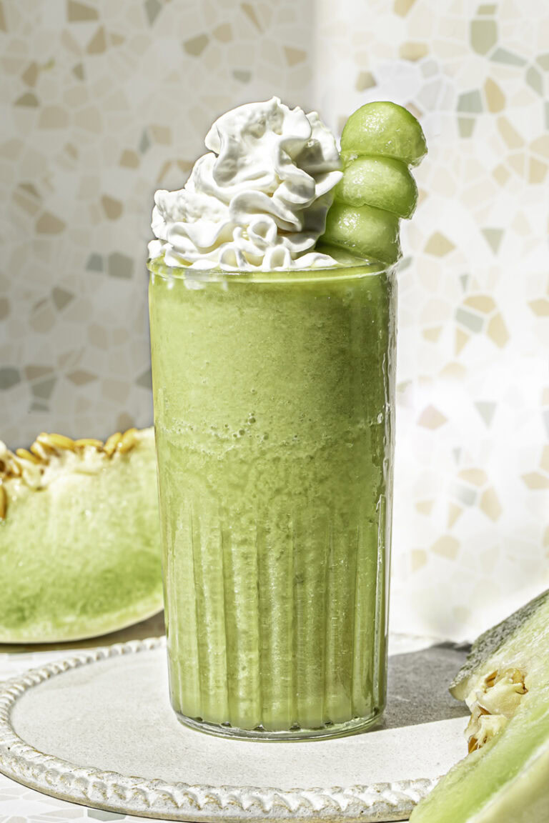 a glass of honeydew green tea topped with whipped cream and a skewer of honeydew balls.