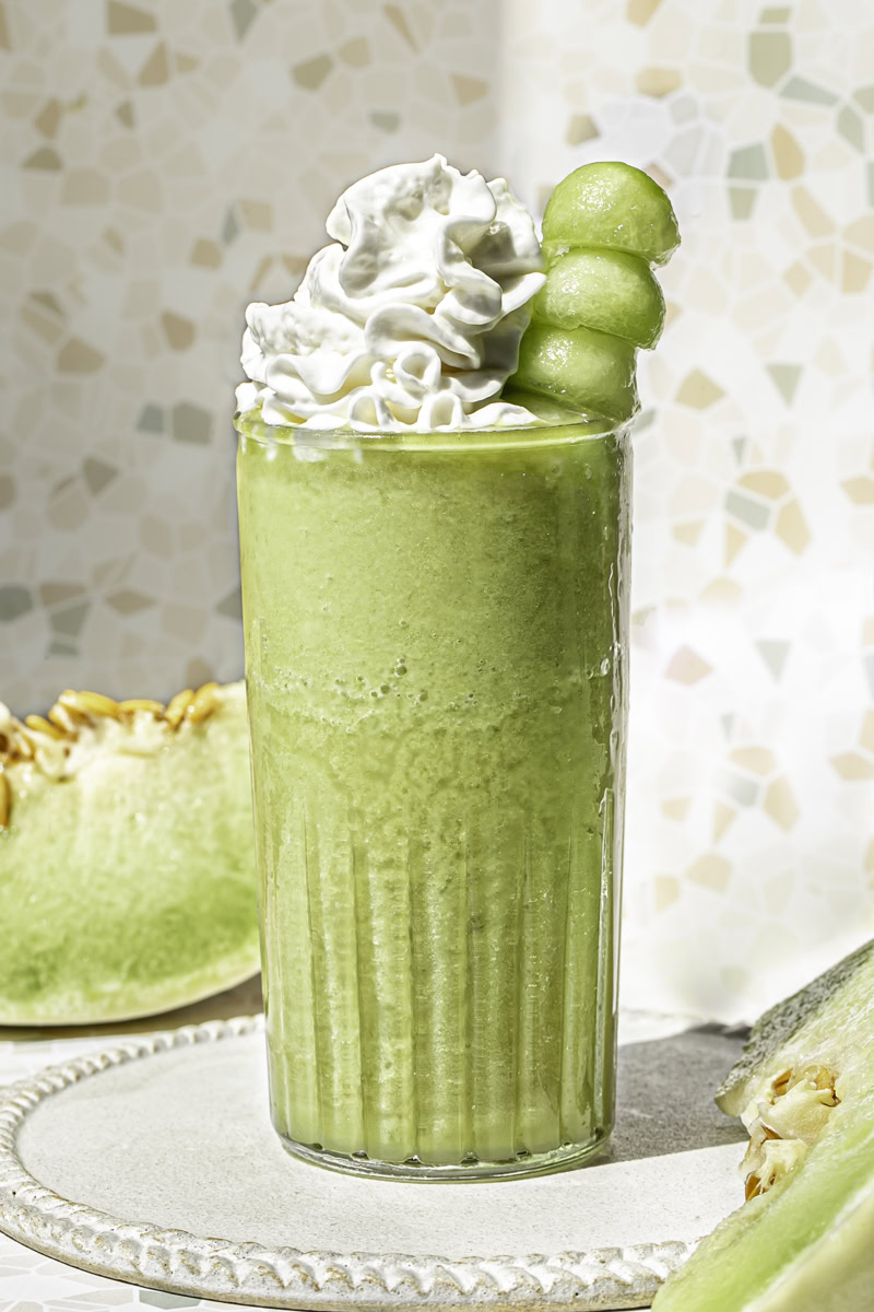 a glass of honeydew green tea topped with whipped cream and a skewer of honeydew balls.