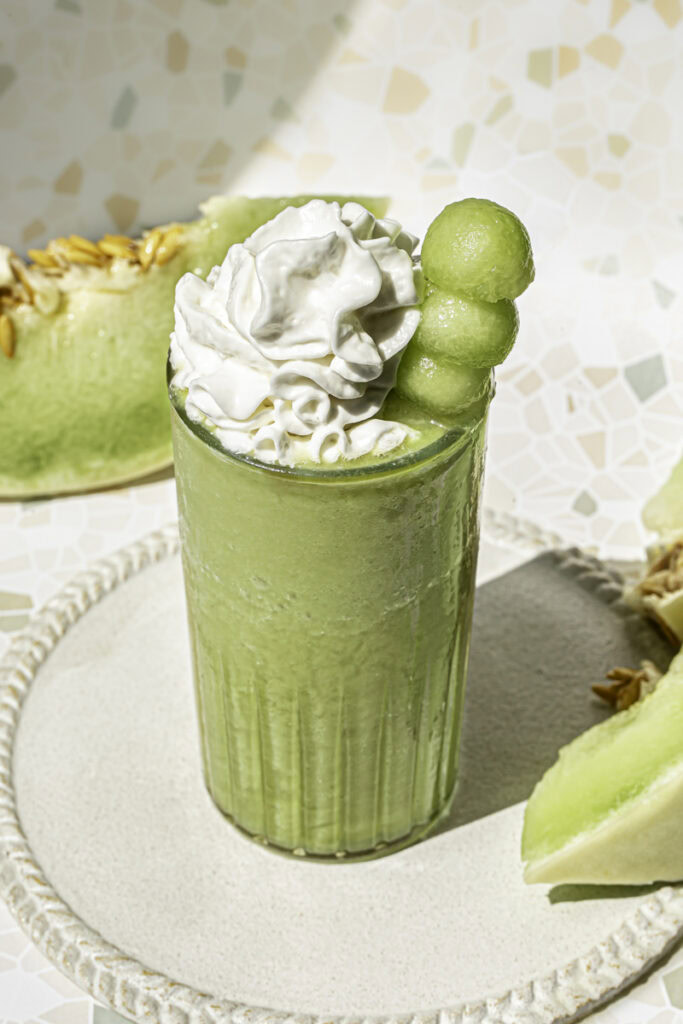 3/4 shot of a glass of honeydew green tea topped with whipped cream and a skewer of honeydew balls and honeydew slices in the background.
