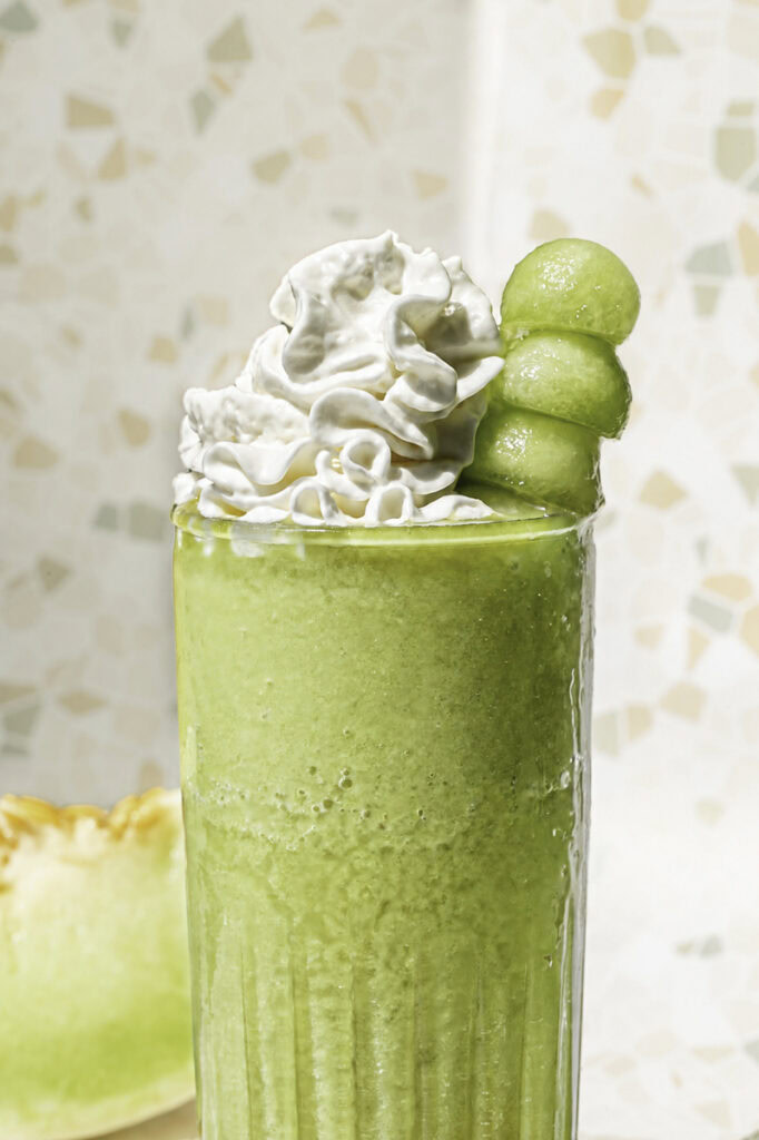 close up of a glass of honeydew green tea topped with whipped cream and a skewer of honeydew balls.