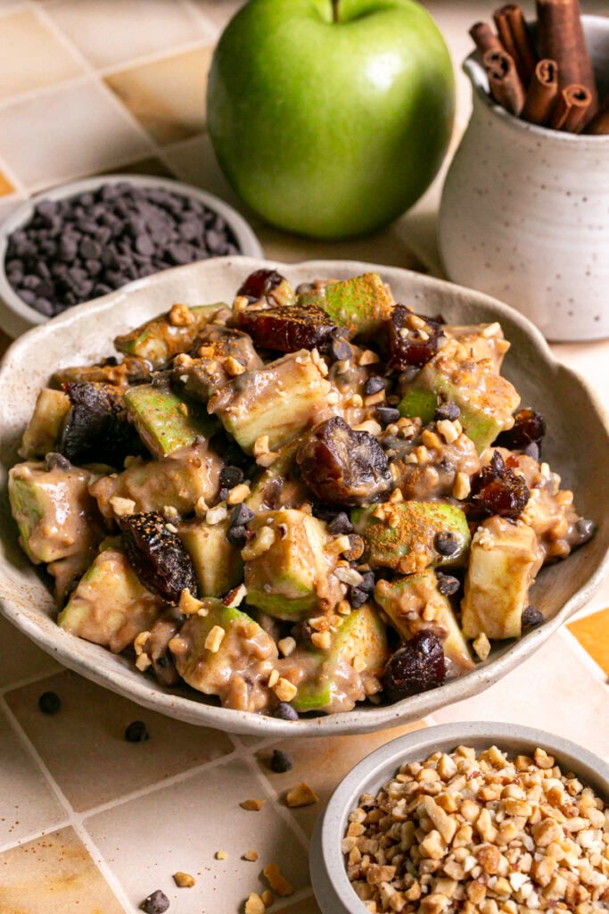 3/4 shot of caramel apple date salad in a bowl with two small bowls filled with chocolate chips and chopped nuts, a green apple, and a jar of cinnamon sticks.