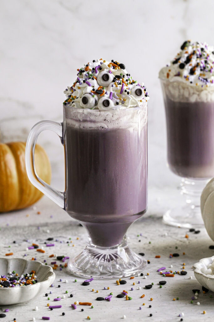 two glasses of Purple Halloween White Hot Chocolate topped with whipped cream, halloween sprinkles and candy eyeballs.
