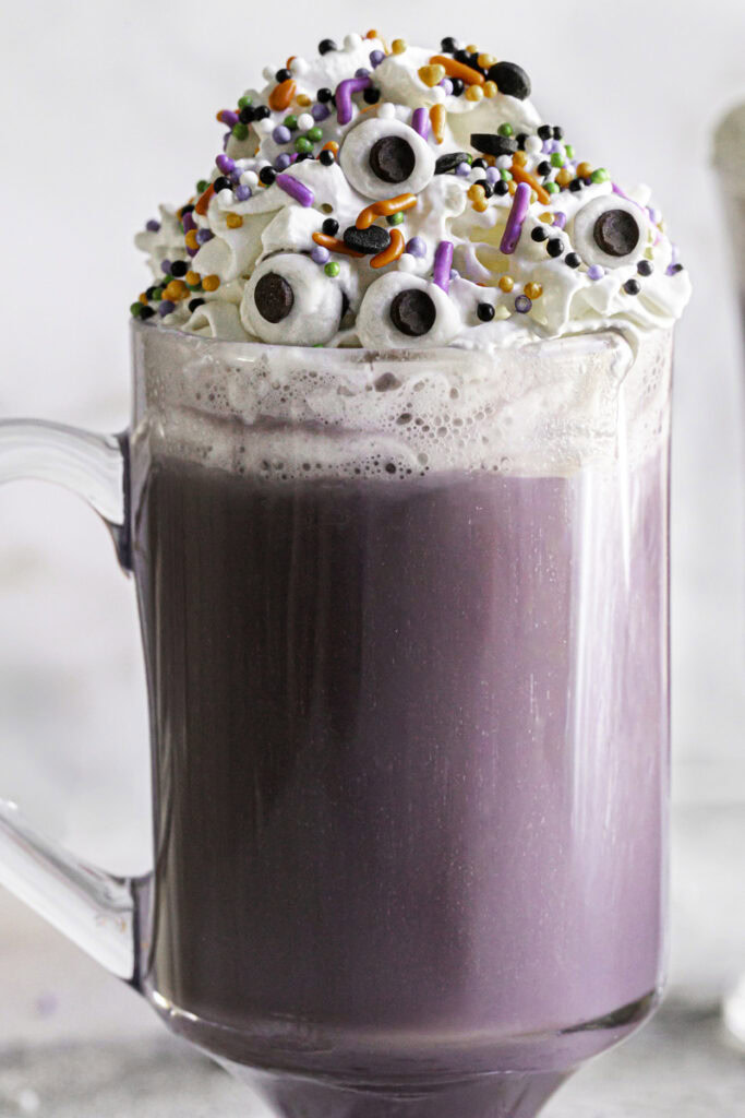 close up shot of a glass of Purple Halloween White Hot Chocolate topped with whipped cream, halloween sprinkles and candy eyeballs.