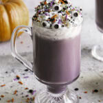 a glass of Purple Halloween White Hot Chocolate topped with whipped cream, halloween sprinkles and candy eyeballs.