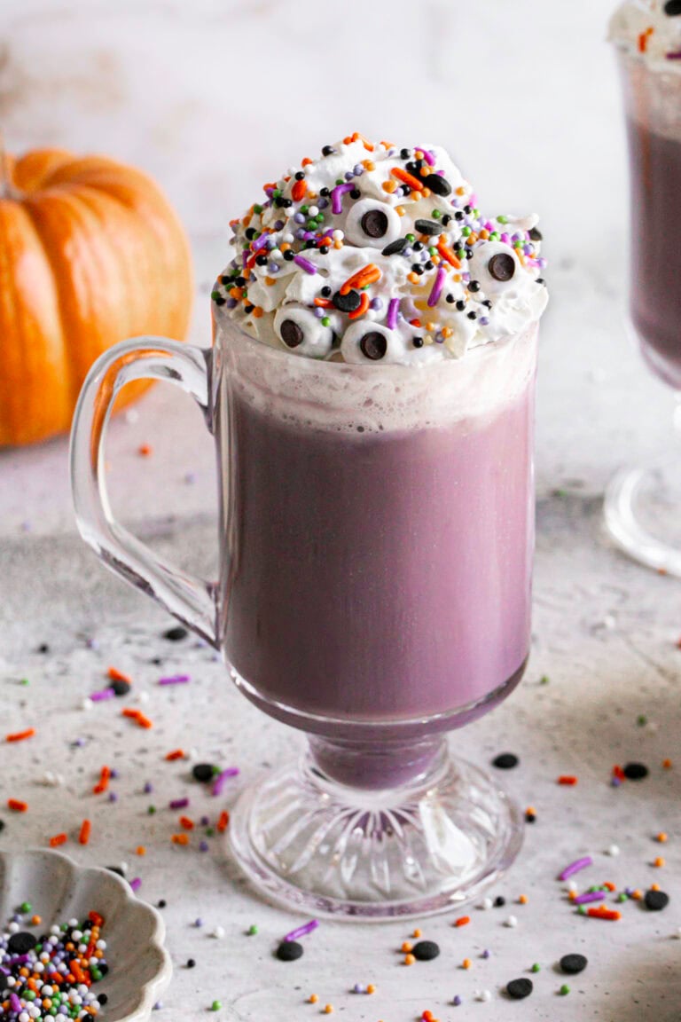 a glass of Purple Halloween White Hot Chocolate topped with whipped cream, halloween sprinkles and candy eyeballs.