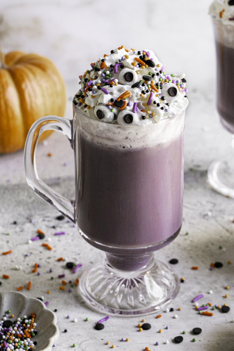 a glass of Purple Halloween White Hot Chocolate topped with whipped cream, halloween sprinkles and candy eyeballs.