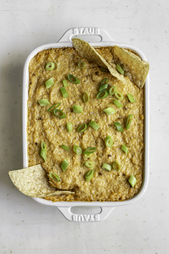 baked buffalo cauliflower dip topped with sliced green onions and tortilla chips in the corner of the dip.