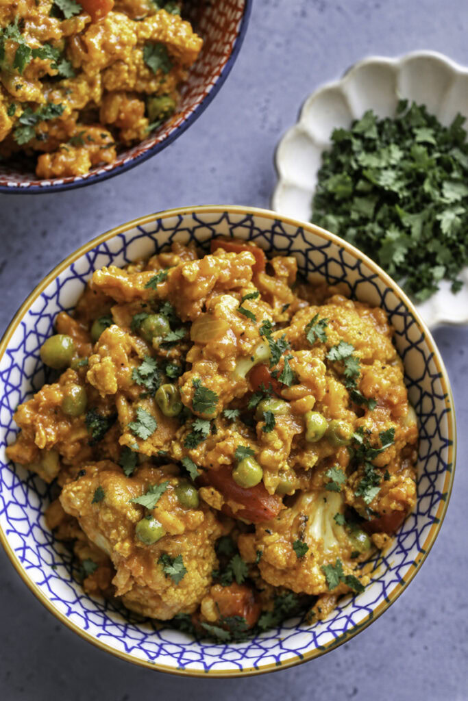 two bowls of Curried Cauliflower with tofu, red bell pepper, and peas and a small bowl of chopped cilantro