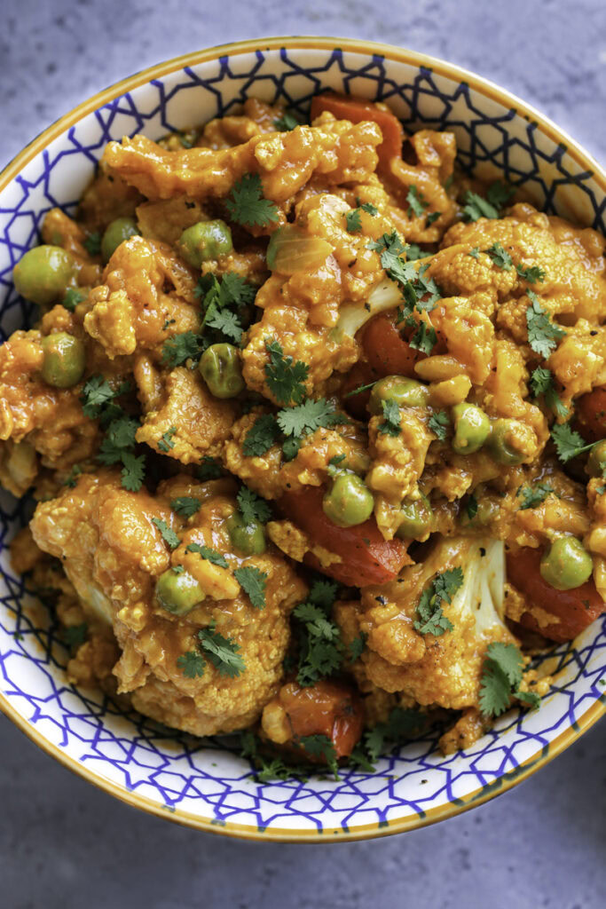 Curried cauliflower with tofu, peas, and red bell peppers in a decorative bowl.