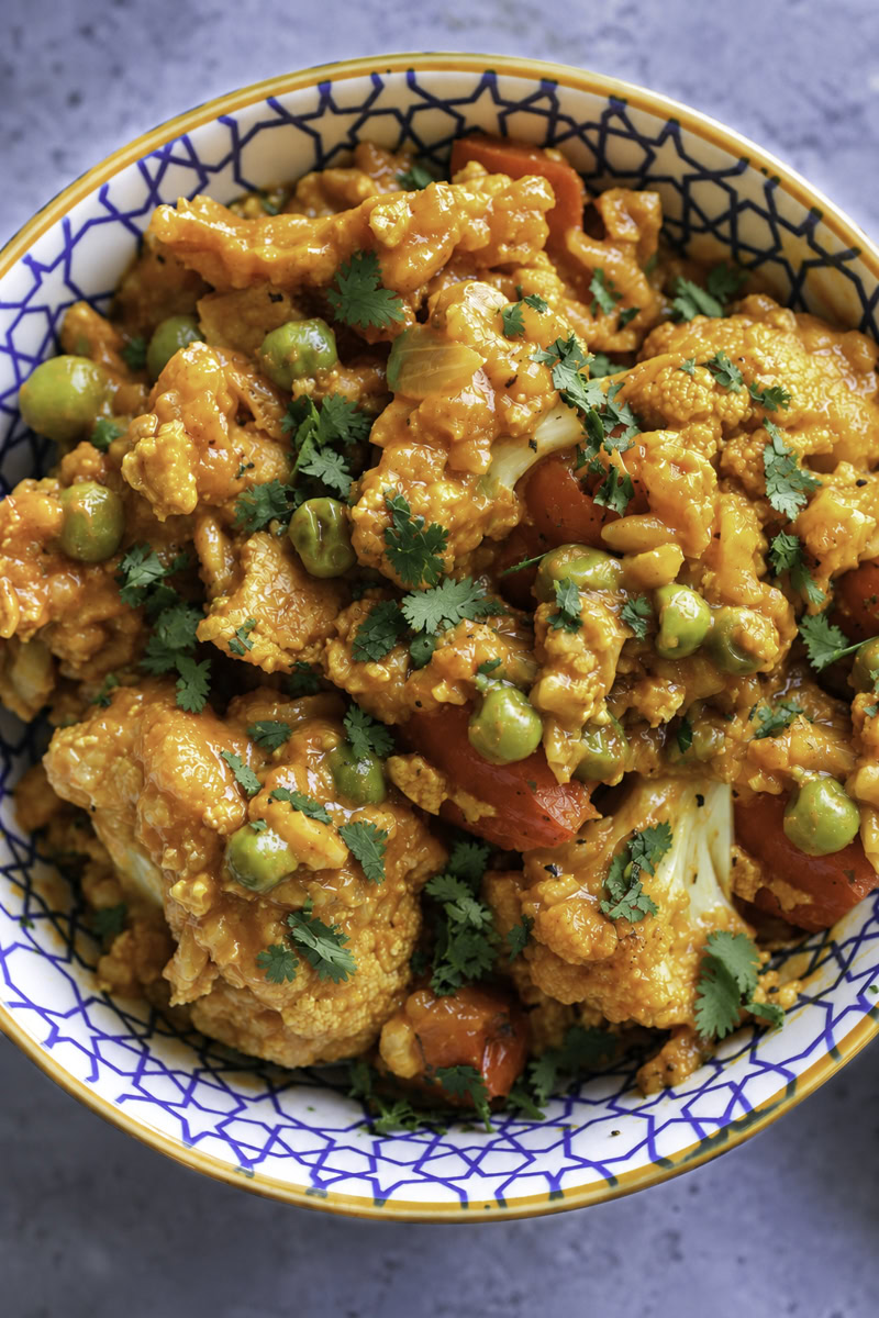 Curried cauliflower with tofu, peas, and red bell peppers in a decorative bowl.