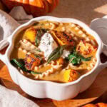 a pumpkin shaped white ceramic bowl filled with creamy roasted pumpkin lasagna soup, with two mini orange pumpkins, a napkin.