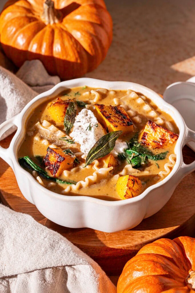 a pumpkin shaped white ceramic bowl filled with creamy roasted pumpkin lasagna soup, with two mini orange pumpkins, a napkin.